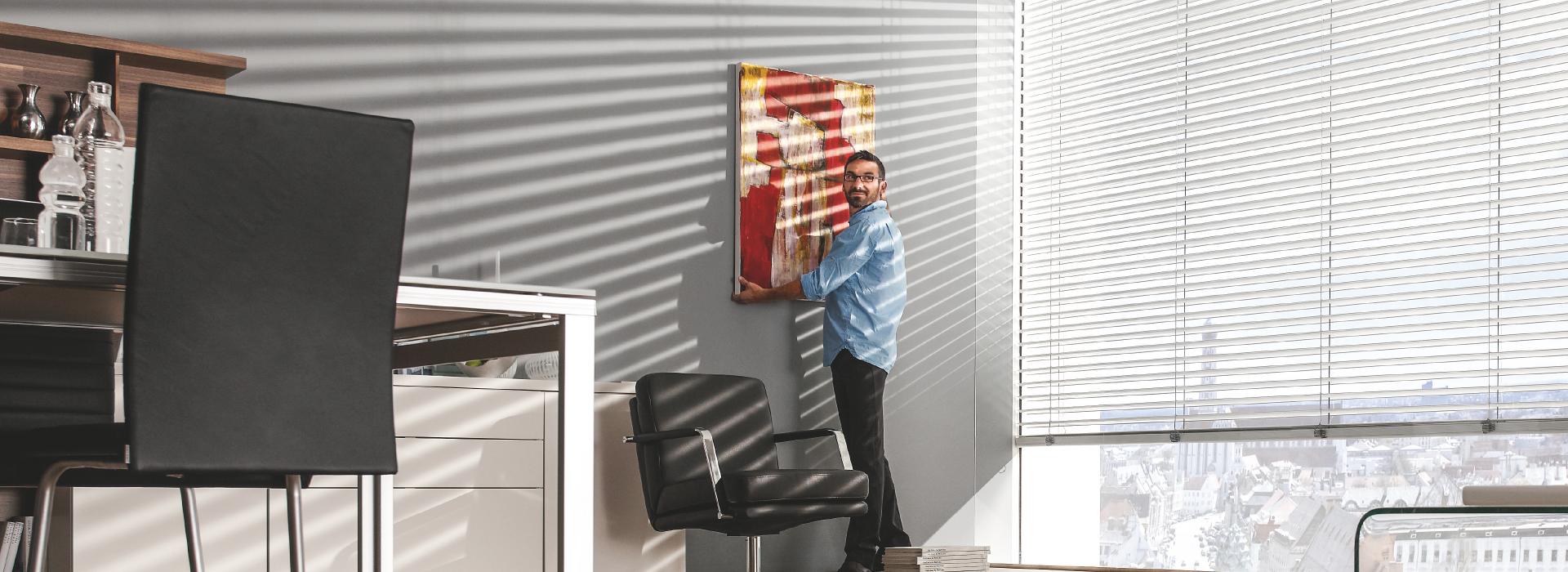 Man hangs up painting in office, external venetian blinds are tilted